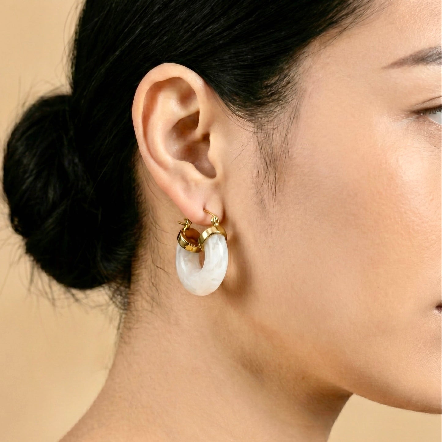 Close-up of a person wearing gold hoop earrings with white stones on a beige background